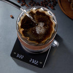 A clear glass mug with a wooden handle sits on a black digital scale displaying time and temperature. Hot water is being poured into a paper coffee filter filled with coffee inside the mug.