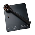 A black digital coffee scale with a matching scoop filled with coffee beans, shown on a plain white background.