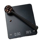 A black digital coffee scale with a matching scoop filled with coffee beans, shown on a plain white background.