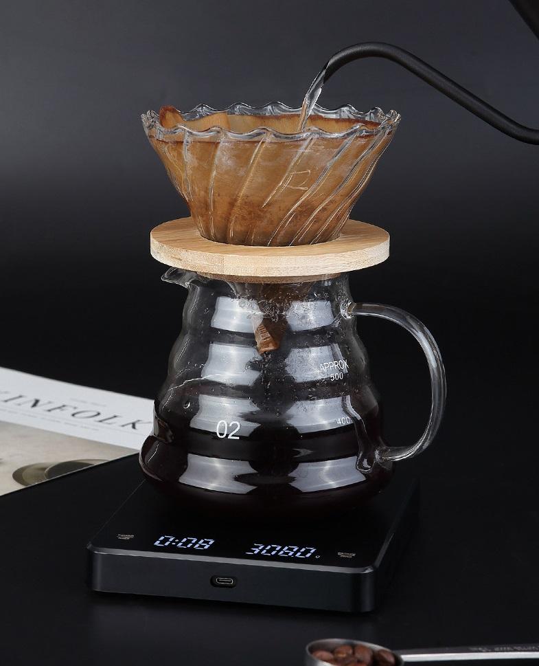 A glass pour-over coffee maker setup on a black digital scale, with coffee dripping from a fluted glass cone filter into a glass carafe, against a dark background.