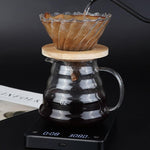 A glass pour-over coffee maker setup on a black digital scale, with coffee dripping from a fluted glass cone filter into a glass carafe, against a dark background.